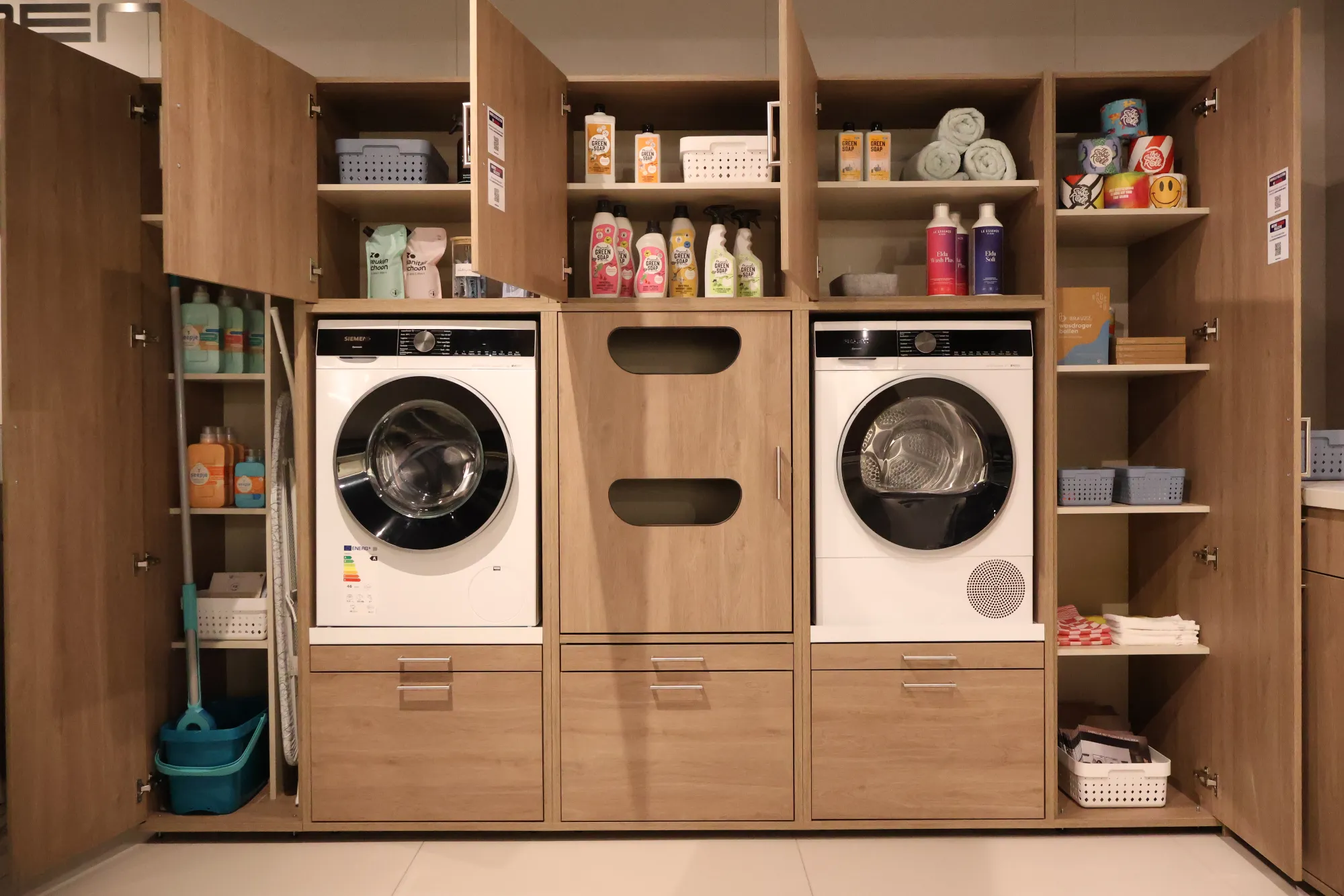 Geöffnete Washtower Schranklösung in Holzoptik mit Waschmaschine, Tumbler und Waschmitteln